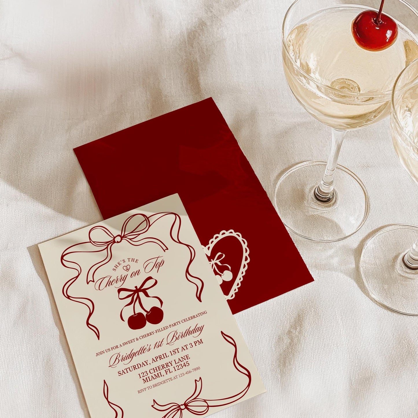May include: A cream colored invitation with red and white accents. The invitation is for a birthday party and has the text 'She's the Cherry on Top' and 'Bridgette's 1st Birthday' with details about the party. The invitation is sitting on a white surface with a red envelope and two glasses of champagne with cherries on top.